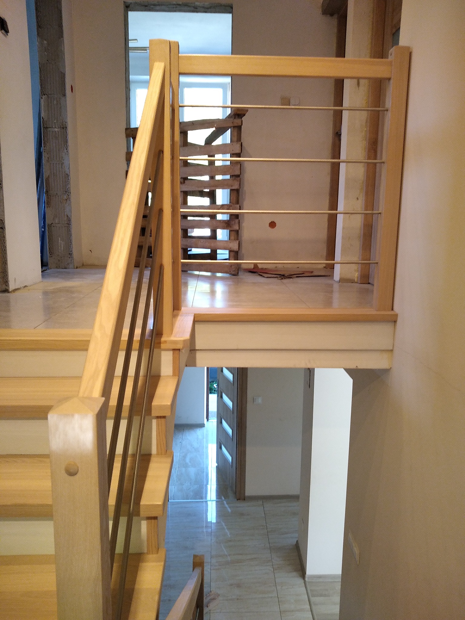 Jasne drewniane schody z metalowymi barierkami w nowym domu, widok z góry na dół, fragment schodów tymczasowych w tle.