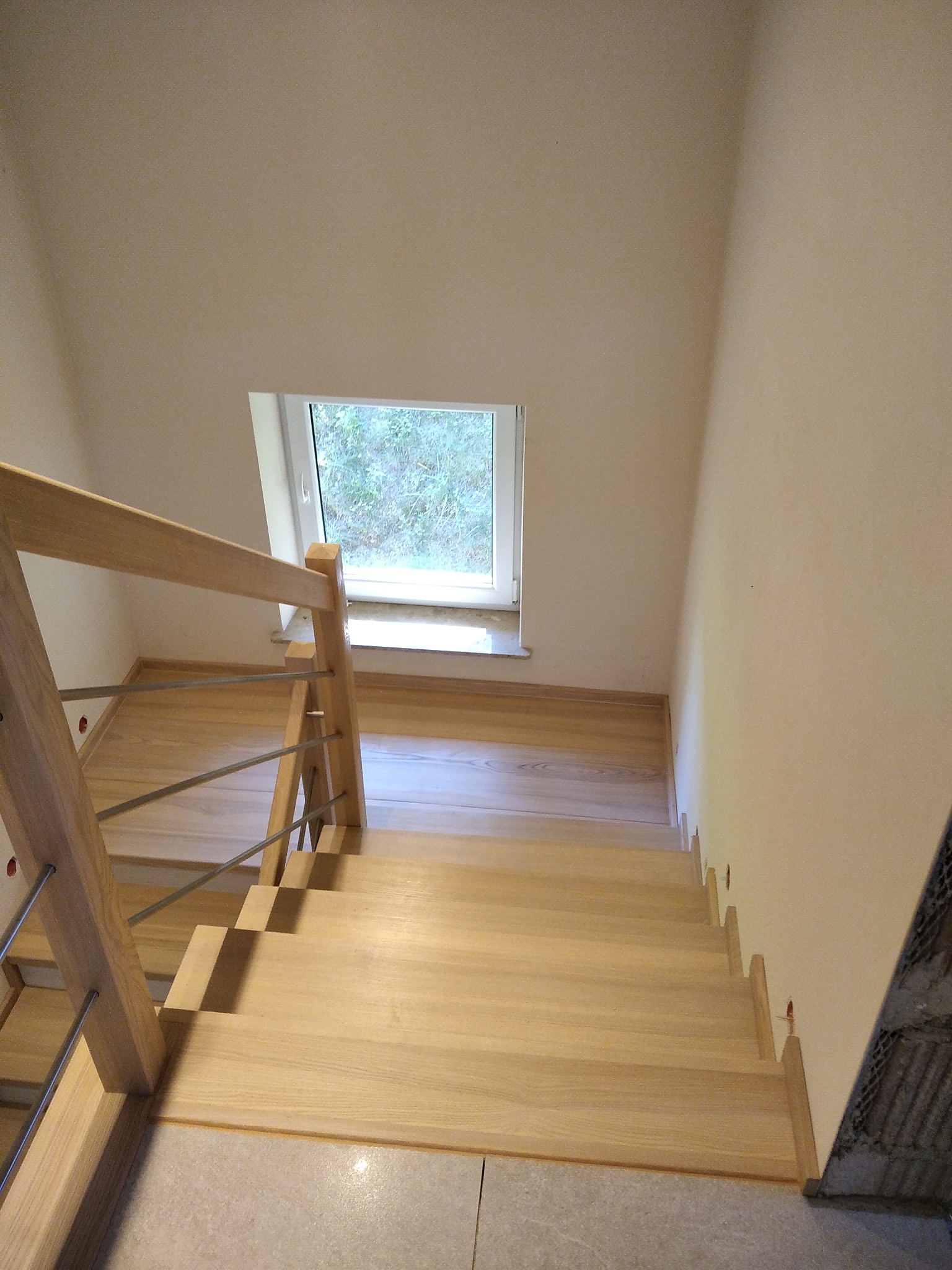 Jasne drewniane schody z metalowymi poręczami, widok z góry, małe okno na półpiętrze, fragment ściany w trakcie wykończenia.