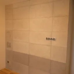 Ściana wyłożona beżowymi i szarymi płytkami, z widocznymi gniazdkami elektrycznymi i fragmentem drewnianej podłogi.