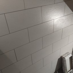 Usługi remontowe i wykończenia wnętrz - Ściana wyłożona podłużnymi, matowymi płytkami w odcieniach szarości, ułożonymi poziomo. W rogu widoczny fragment opakowania płytek.