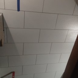 Usługi remontowe i wykończenia wnętrz - Ułożone białe, prostokątne płytki na ścianie, z widoczną niebieską taśmą malarską i poziomicą. Widoczny fragment konstrukcji drewnianej.