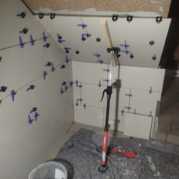 Usługi remontowe i wykończenia wnętrz - Białe płytki na ścianie pod skosem, z niebieskimi krzyżykami dystansowymi. Widoczny kubeł z krzyżykami, narzędzia i podpórka do płytek. Praca w toku.