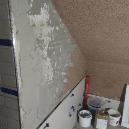 Usługi remontowe i wykończenia wnętrz - Ściana w trakcie kładzenia płytek w łazience na poddaszu. Widoczne narzędzia, poziomica i klej. Częściowo położone płytki z niebieskimi krzyżykami dystansowymi.