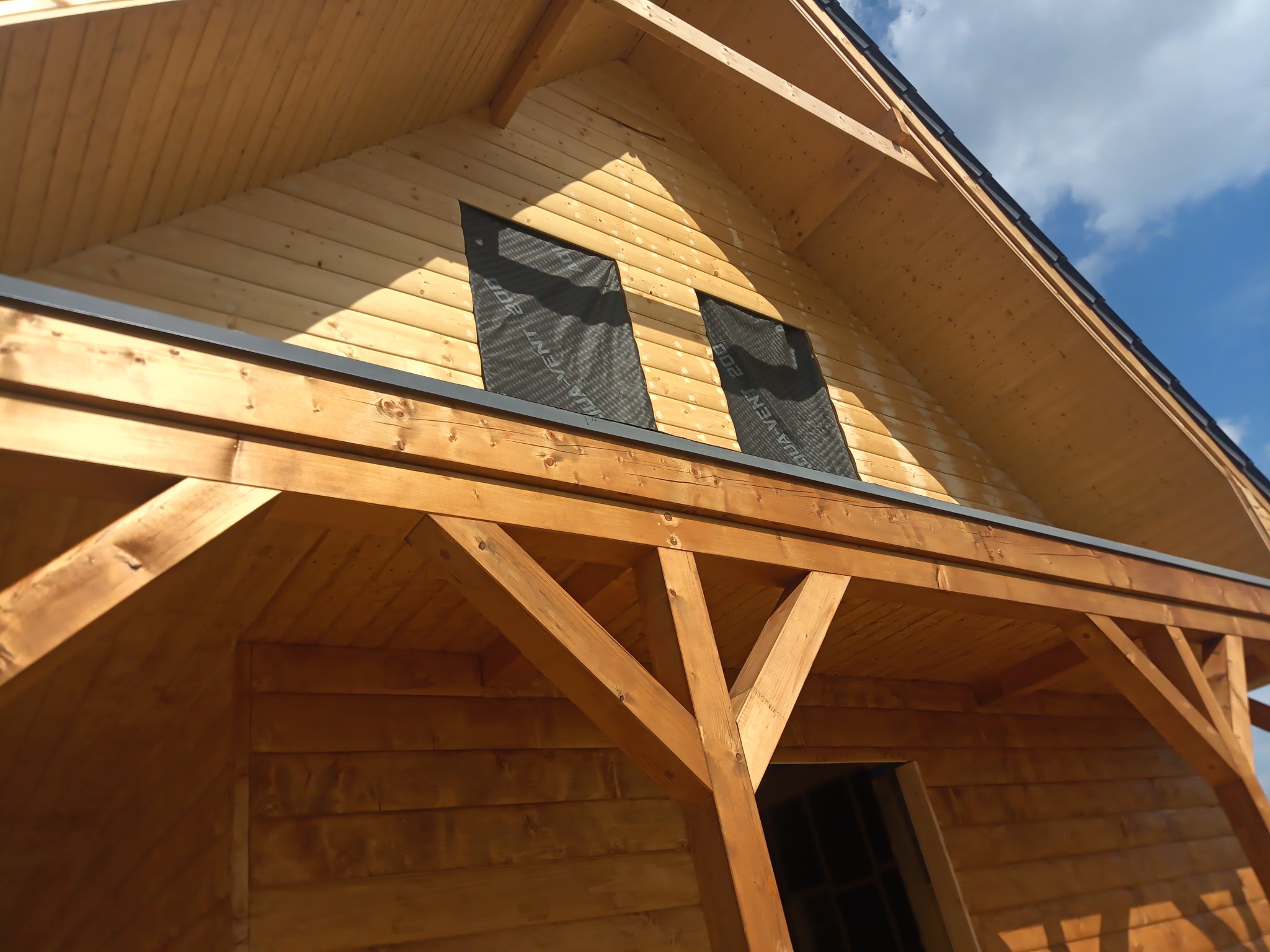 Fragment konstrukcji domu z drewna, widoczne zabezpieczone otwory okienne folią paroizolacyjną i elementy więźby dachowej.