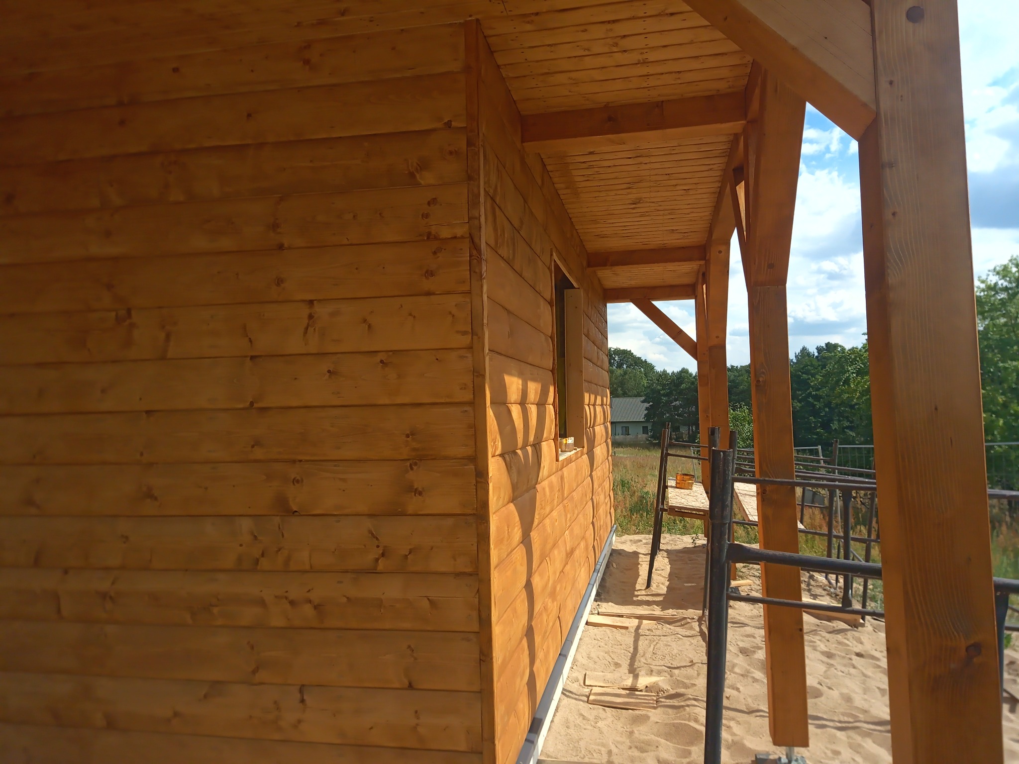 Surowa drewniana konstrukcja domu z widoczną elewacją z desek, odkrytą werandą i niezabudowanym oknem, stojąca na piaszczystym podłożu, z drabina w tle.