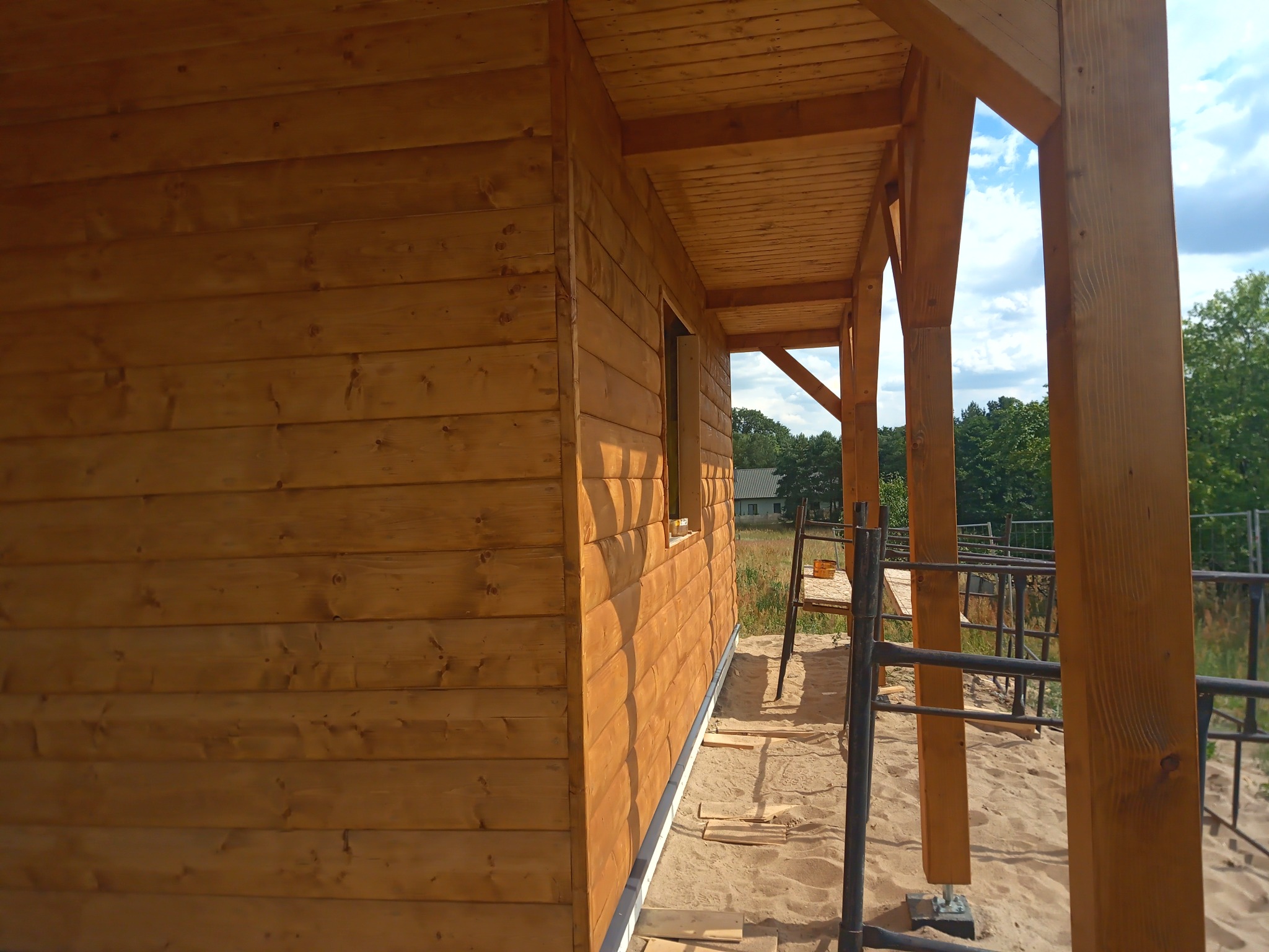 Surowa, drewniana elewacja domu w trakcie budowy, widoczne drewniane belki konstrukcyjne i fragment okna, w tle drabina i zieleń.