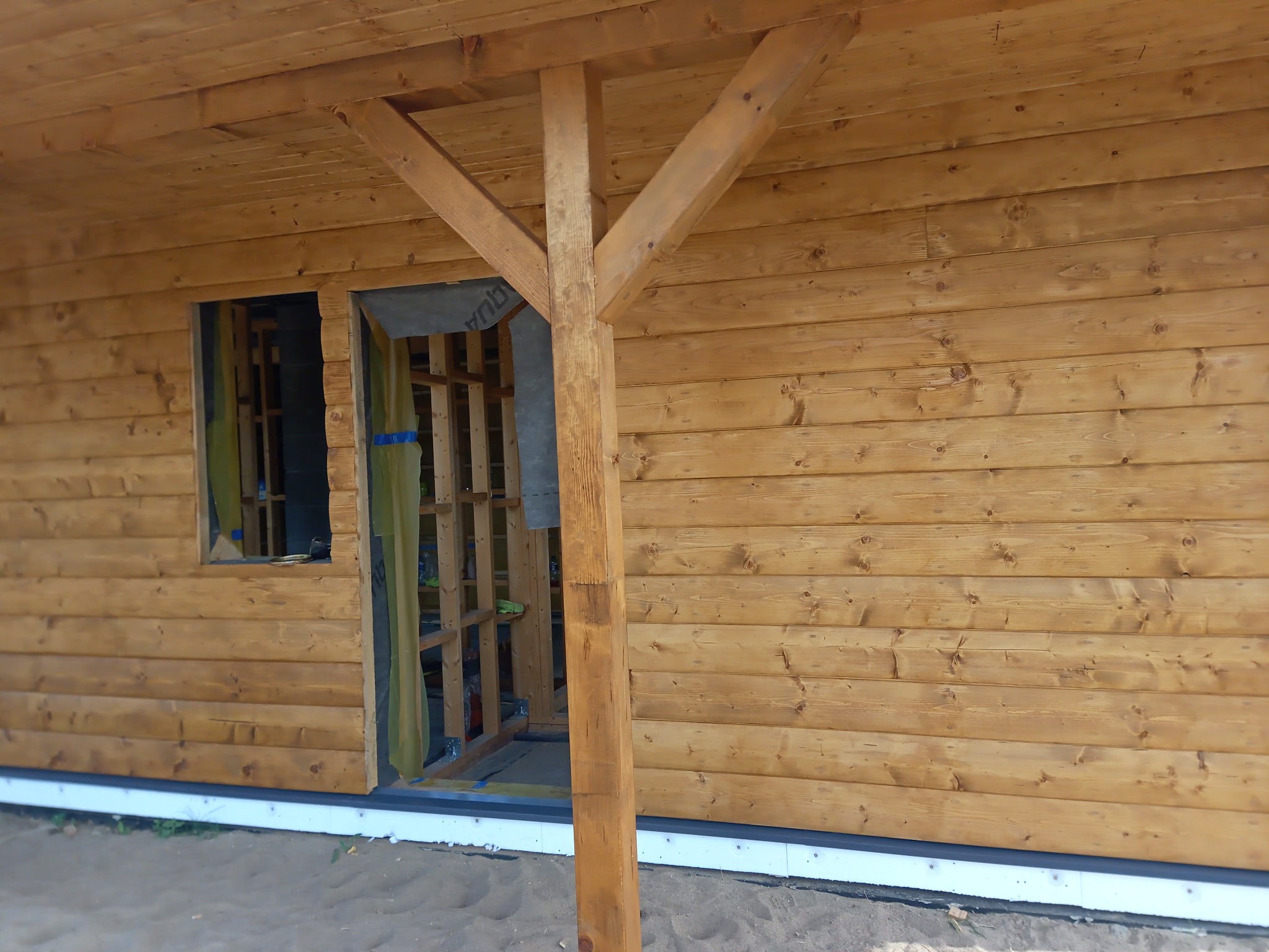 Surowa drewniana konstrukcja domu w trakcie budowy, widoczne elementy szkieletu i izolacji, z zewnątrz obłożona deskami, wsparta na drewnianym słupie.