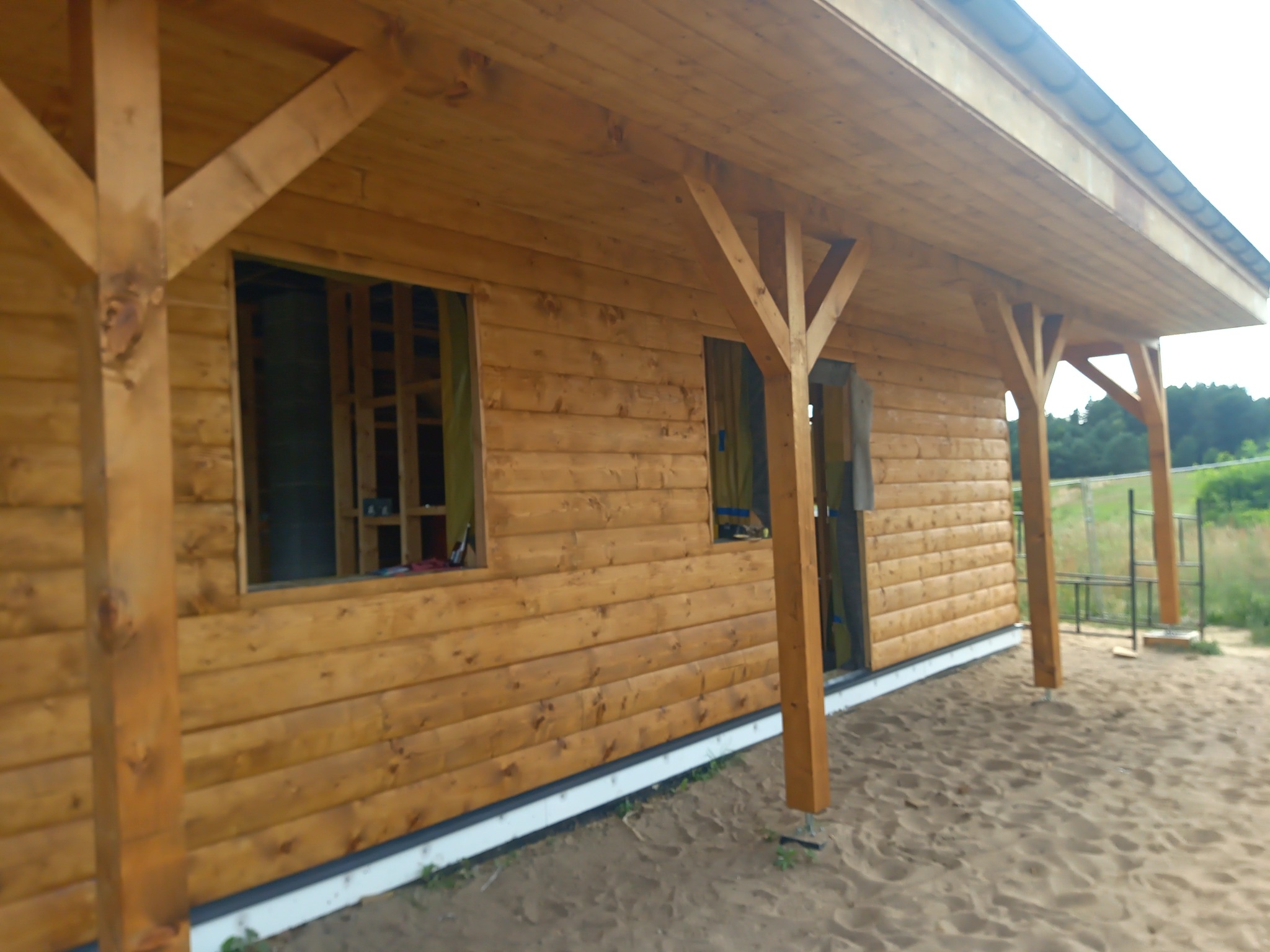 Surowy drewniany budynek z niezabezpieczonymi otworami okiennymi i drzwiowymi, z widoczną konstrukcją dachu i podporami, na piaszczystym terenie.