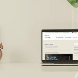 Laptop wyświetlający stronę internetową Marcina Rosińskiego na biurku z dekoracjami: miedziany budzik retro, biała ramka i zielony liść monstery.