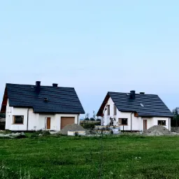 Dwa identyczne, nowe domy jednorodzinne z białymi elewacjami i ciemnymi dachami, widoczne stosy piasku i materiałów budowlanych przed budynkami, sugerujące trwające prace wykończeniowe.