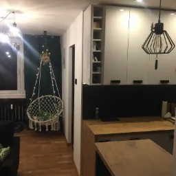 Wnętrze mieszkania z drewnianą podłogą i nowoczesną kuchnią z białymi szafkami, w tle okno z zasłoną i wiszący fotel hamakowy z dekoracjami.