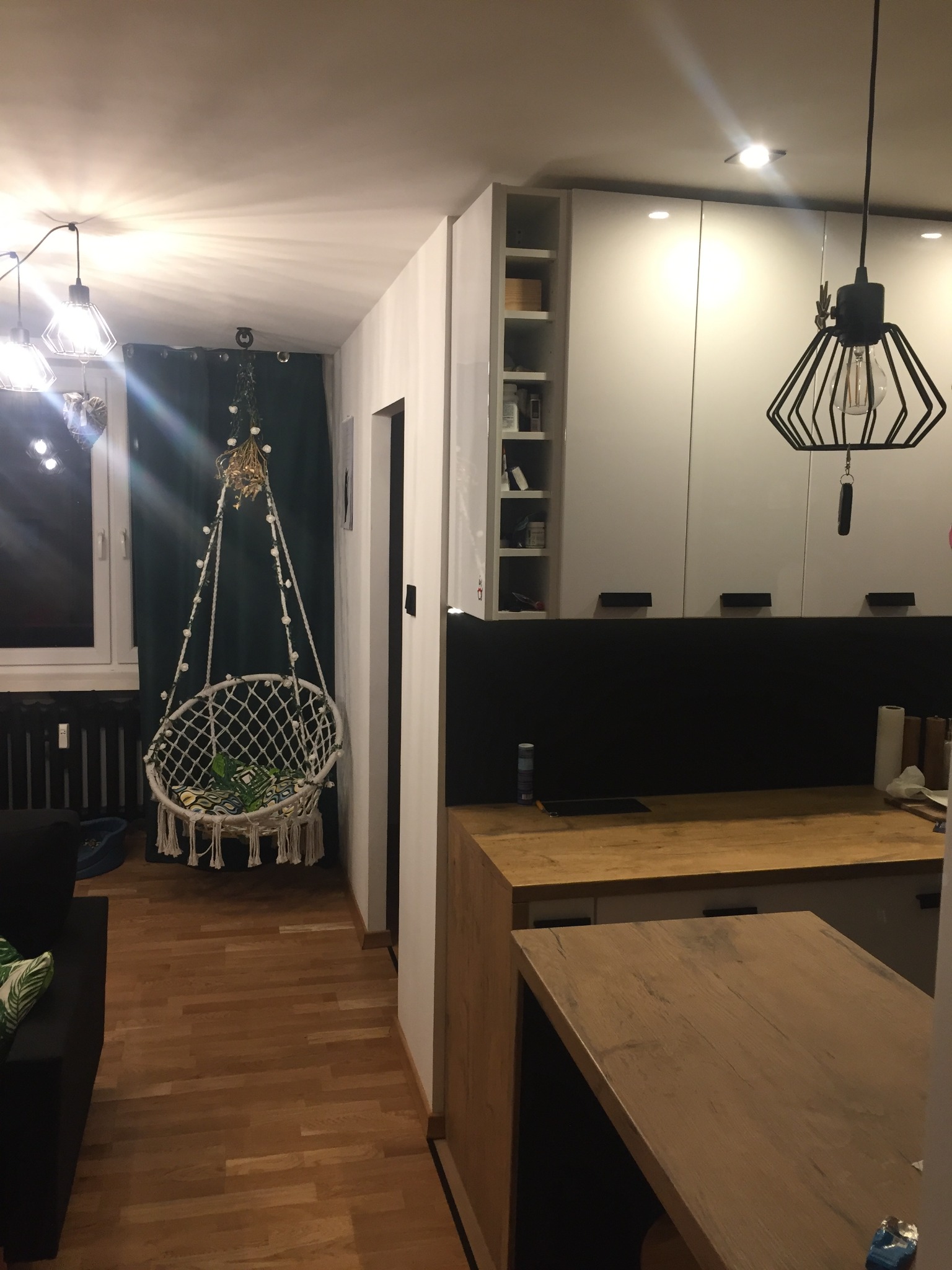 Wnętrze mieszkania z drewnianą podłogą i nowoczesną kuchnią z białymi szafkami, w tle okno z zasłoną i wiszący fotel hamakowy z dekoracjami.