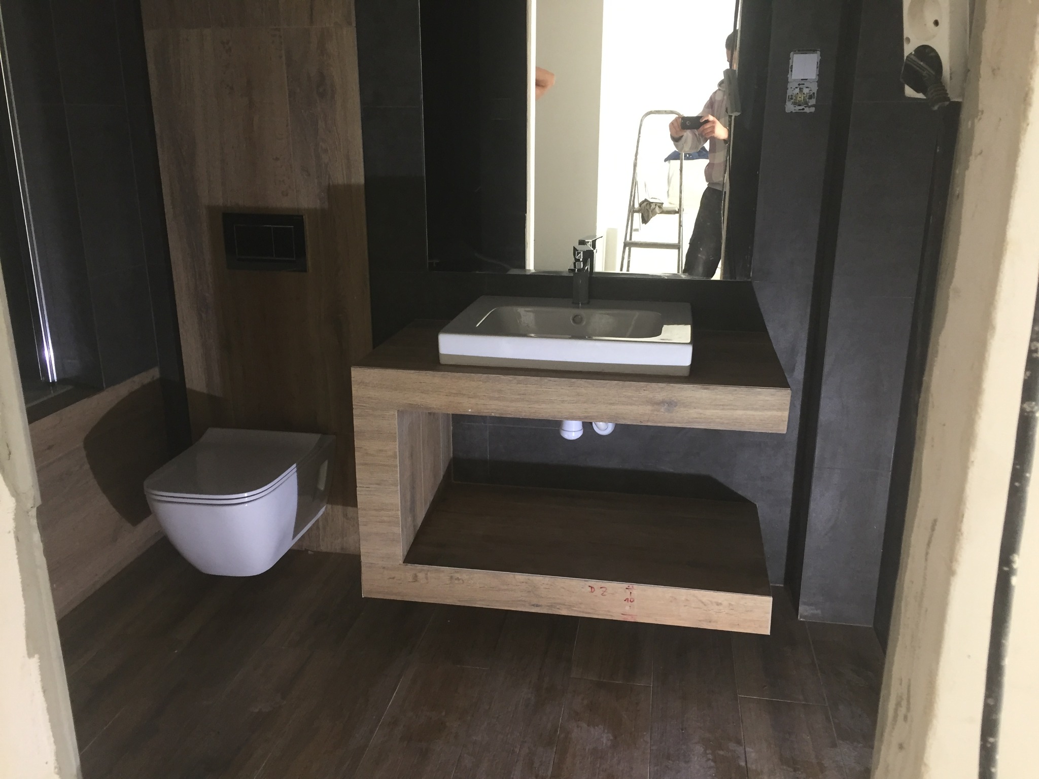 Nowoczesna łazienka w trakcie remontu: podwieszana toaleta, umywalka nablatowa na drewnianym blacie, czarne płytki na ścianach, widoczne odbicie osoby na drabinie w lustrze.