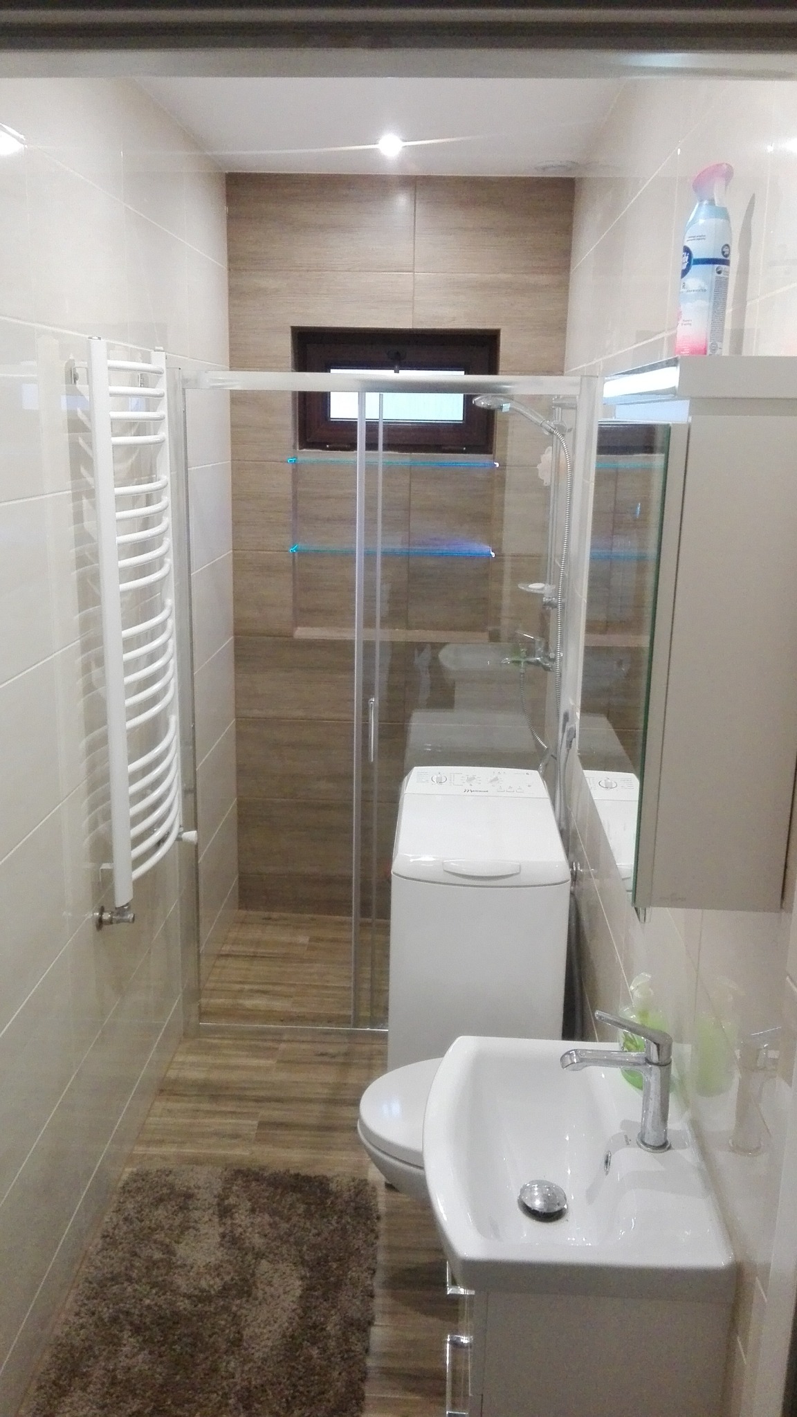 Umeblowana łazienka z kabiną prysznicową, pralką, umywalką z szafką i grzejnikiem drabinkowym, wykończona płytkami w odcieniach beżu i brązu.