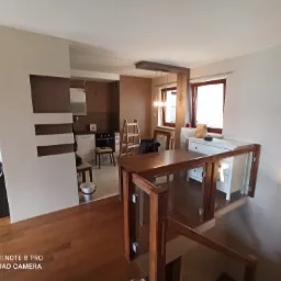 Wnętrze mieszkania po remoncie z widoczną kuchnią, schodami z drewnianą balustradą i szklanym wypełnieniem, oraz ścianą z dekoracyjnymi elementami w odcieniach brązu i beżu.