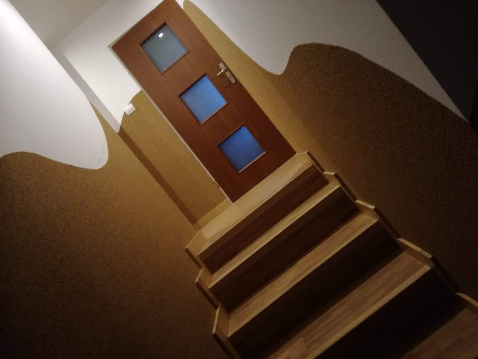 Drewniane schody prowadzące do brązowych drzwi z trzema niebieskimi szybkami, ściany wykończone w odcieniach beżu i bieli.