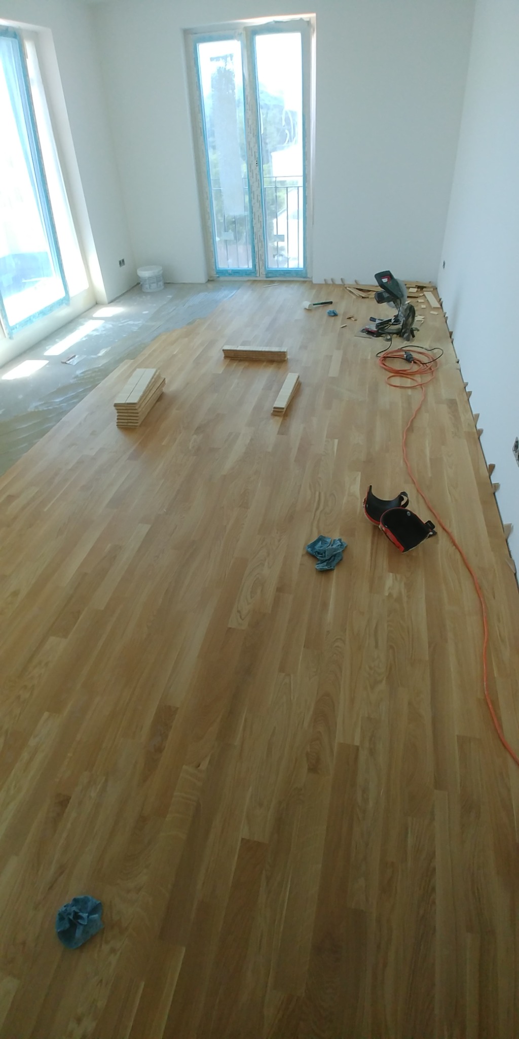 Układanie parkietu w jasnym pomieszczeniu z oknami; widoczny stos desek, piła elektryczna, ochraniacze kolan i szmatka.