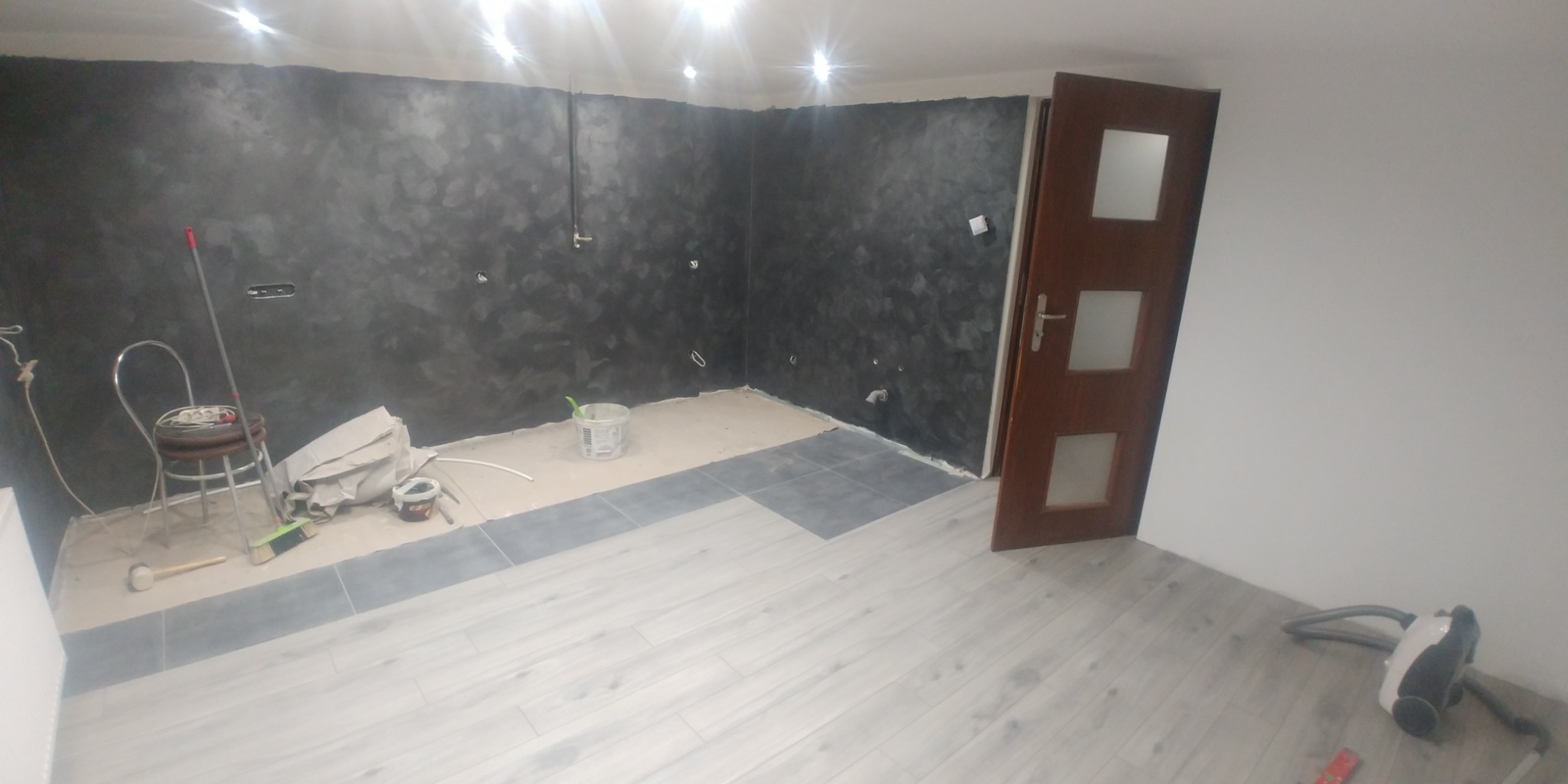Pokój w trakcie remontu: ściany pomalowane na ciemny, wzorzysty kolor, częściowo położona podłoga z paneli i płytek, widoczny stołek, wiadro i narzędzia.