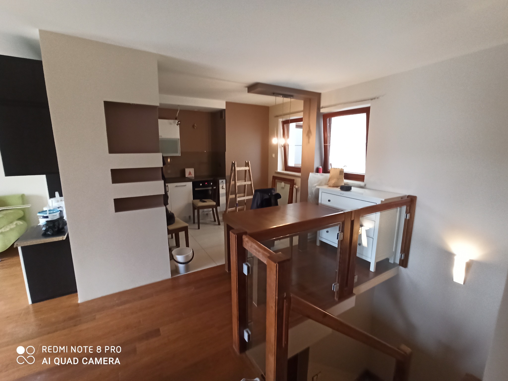 Wnętrze mieszkania po remoncie z widoczną kuchnią, schodami z drewnianą balustradą i szklanym wypełnieniem, oraz ścianą z dekoracyjnymi elementami w odcieniach brązu i beżu.