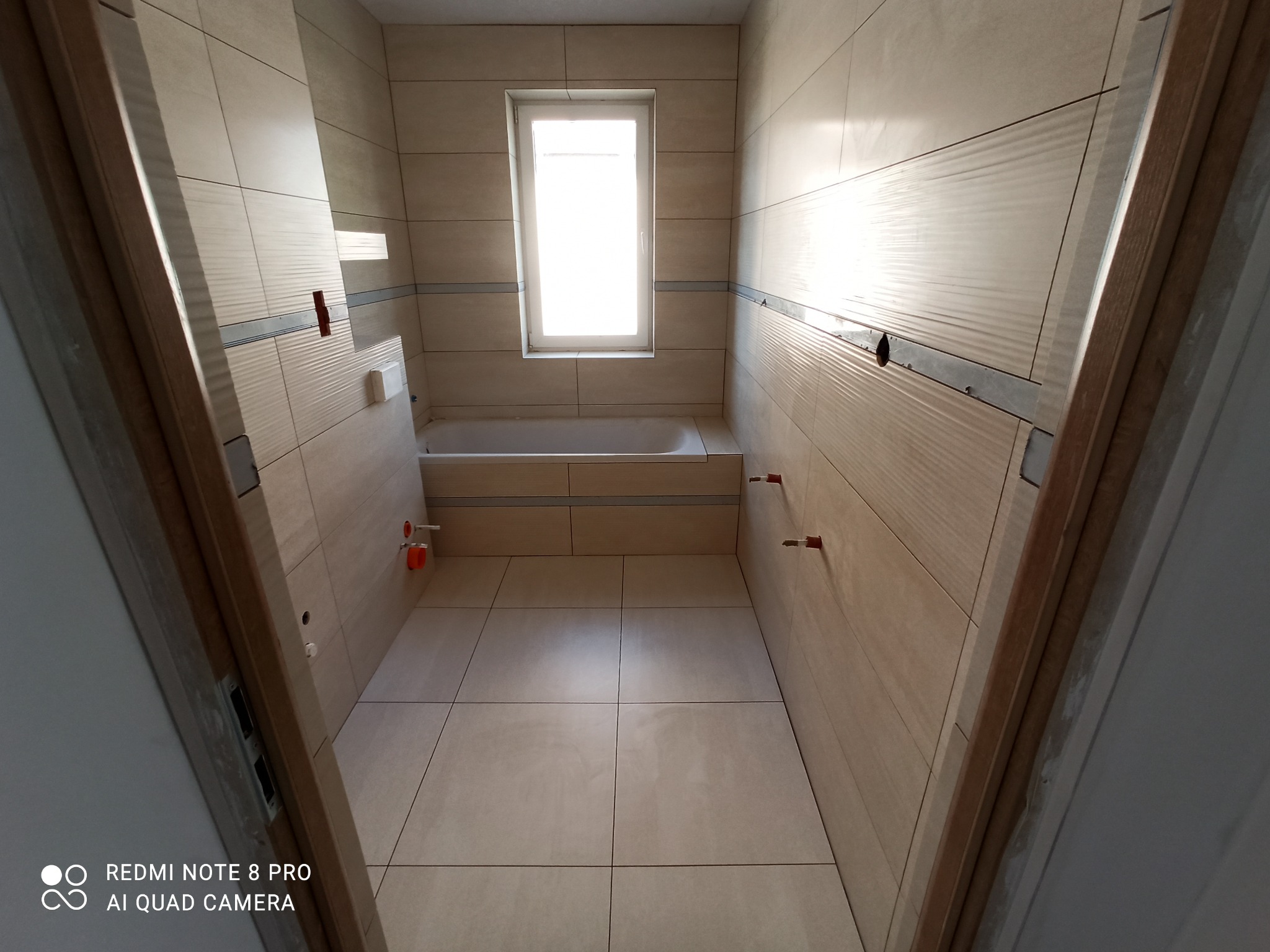 Wnętrze łazienki w trakcie wykańczania, widoczne beżowe płytki na ścianach i podłodze, przygotowane instalacje wodno-kanalizacyjne i okno z widokiem na zewnątrz.