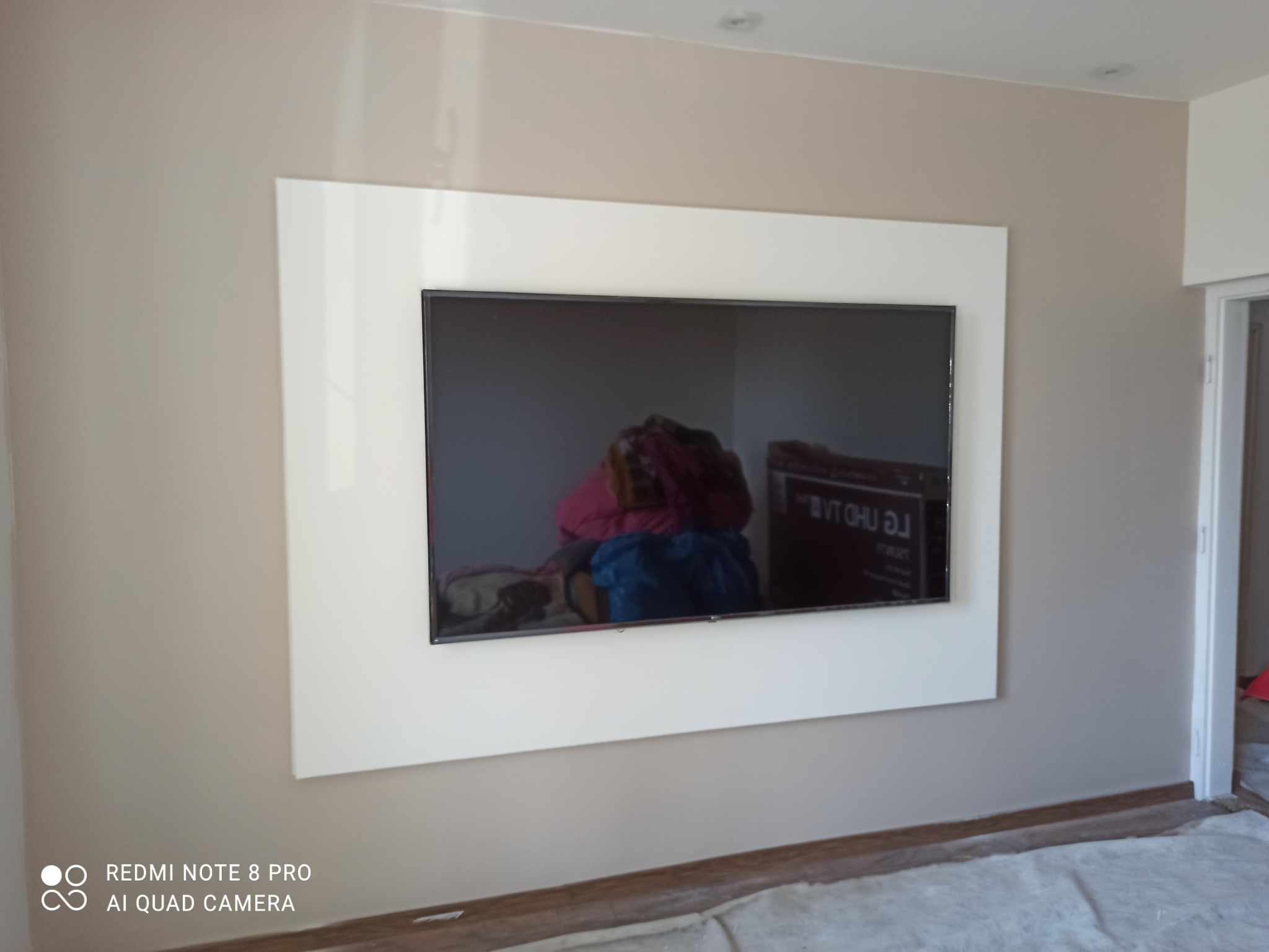 Telewizor zamontowany w białej ramie na beżowej ścianie, podłoga zabezpieczona folią malarską