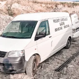 Biały samochód dostawczy z otwartymi drzwiami, na którym widnieje napis 'Projekty i instalacje gazowe' oraz numery telefonów, zaparkowany na placu budowy z widocznymi rurami i drabiną.