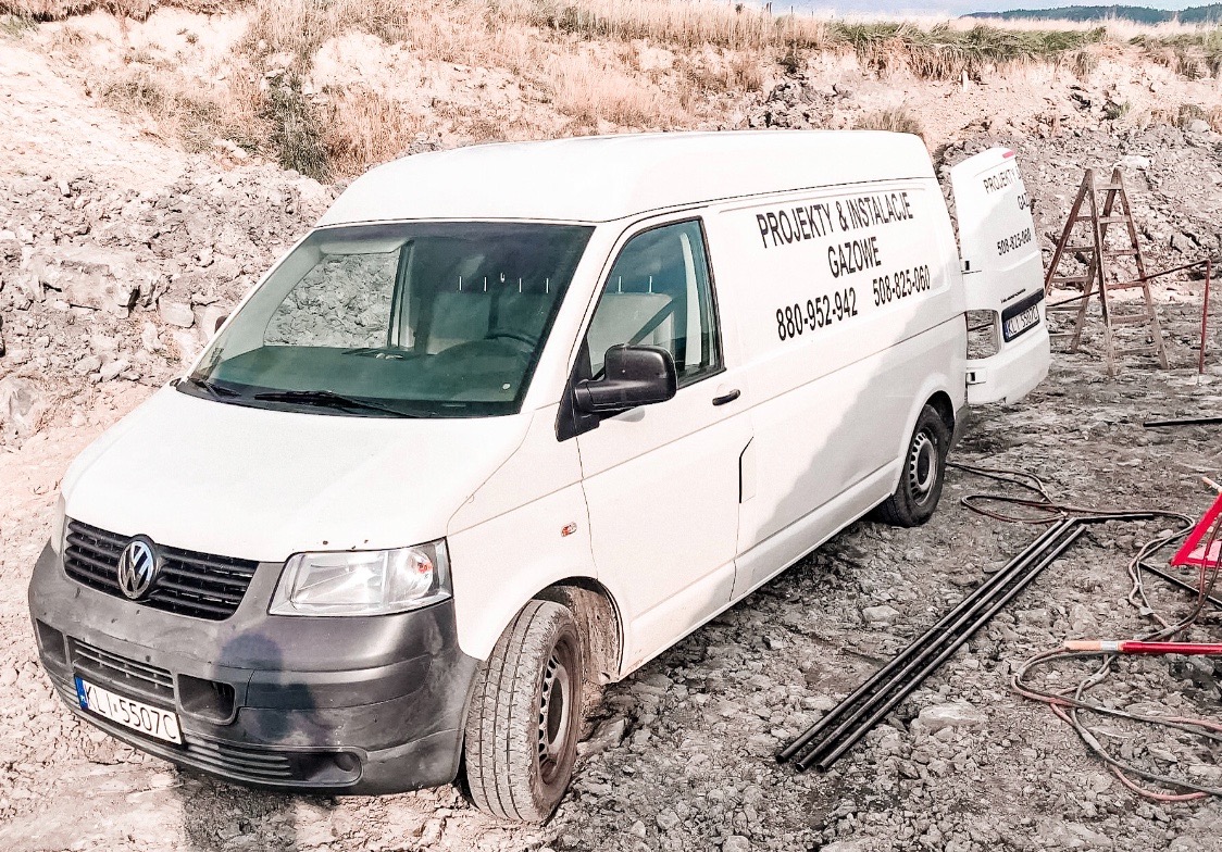 Biały samochód dostawczy z otwartymi drzwiami, na którym widnieje napis 'Projekty i instalacje gazowe' oraz numery telefonów, zaparkowany na placu budowy z widocznymi rurami i drabiną.