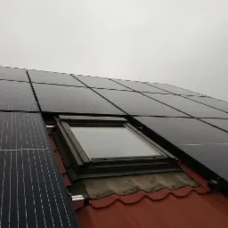 Instalacja fotowoltaiczna na dachu z czerwoną dachówką, z widocznym oknem dachowym i szarym niebem w tle.