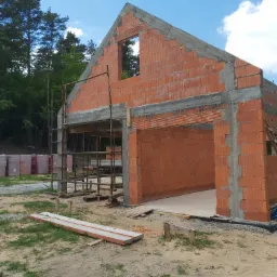 Surowy szkielet domu z czerwonej cegły, widoczne rusztowania i palety z cegłami w tle, błękitne niebo z chmurami.