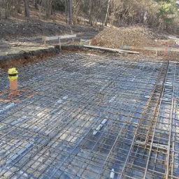 Zbrojenie fundamentów na folii hydroizolacyjnej, z widoczną rurą kanalizacyjną i drewnianymi szalunkami w tle, teren budowy w lesie.