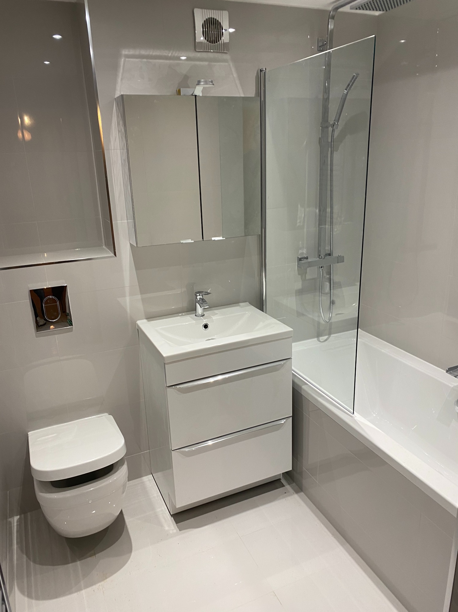 Nowoczesna łazienka z umywalką na szafce z szufladami, podwieszaną toaletą i wanną za szklanym parawanem, wykończona jasnymi płytkami w neutralnym odcieniu szarości.