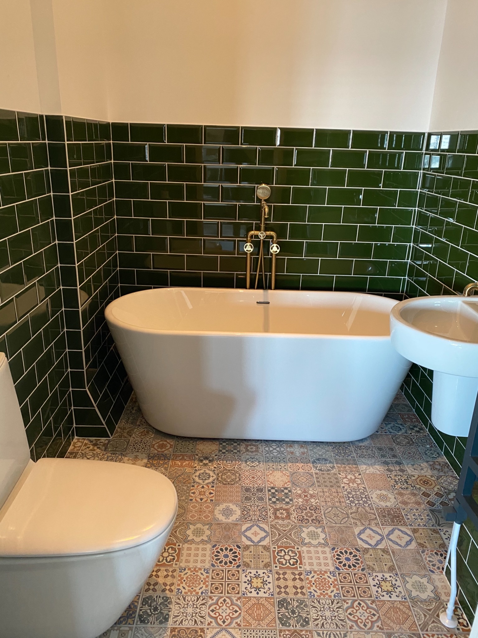 Biała wanna wolnostojąca z mosiężną armaturą na tle ciemnozielonych płytek metra, wzorzysta podłoga z płytek patchworkowych, toaleta i umywalka na postumencie.