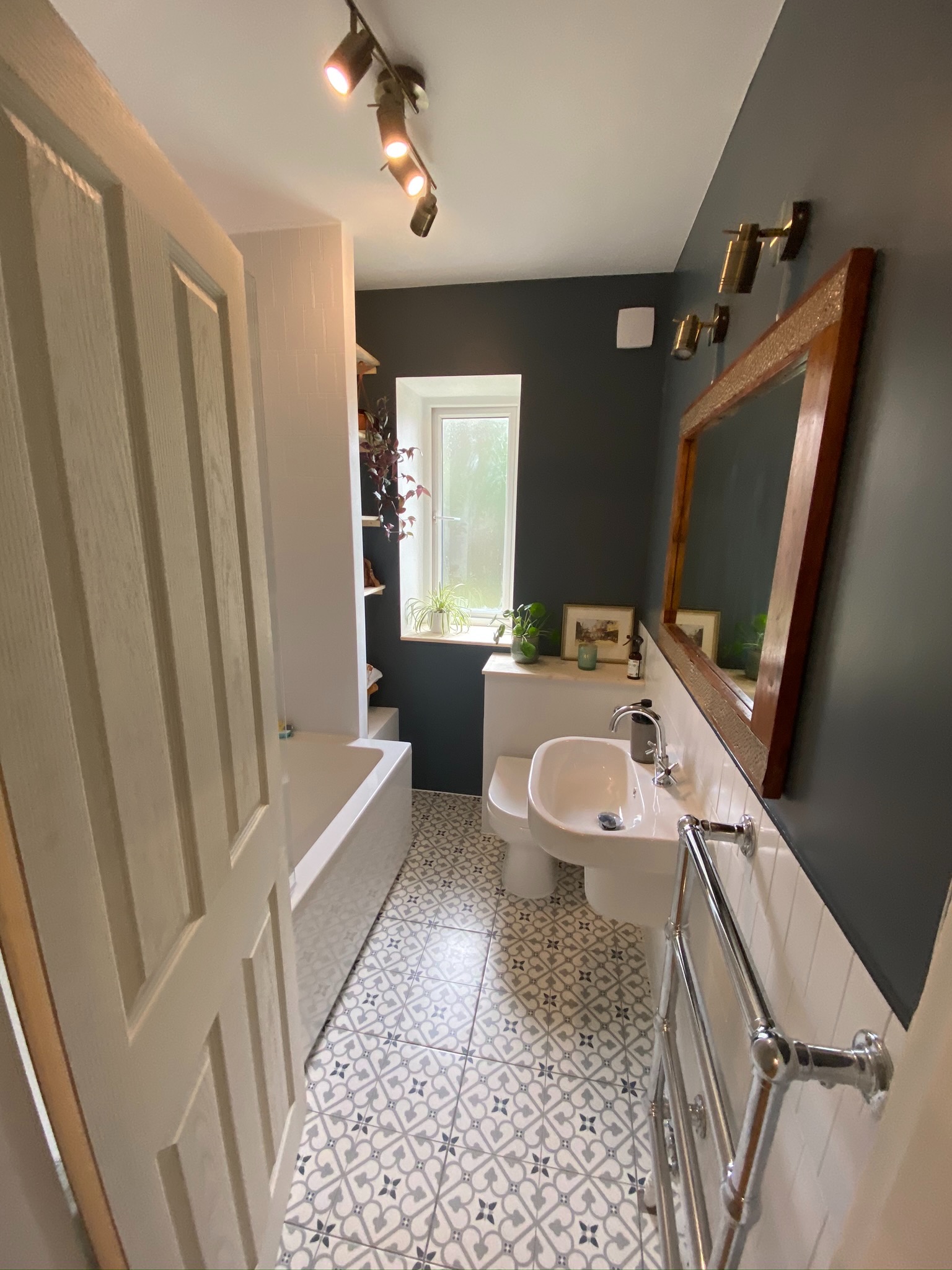 Wąska łazienka z ciemnoszarą ścianą, wzorzystą podłogą, umywalką nablatową i drewnianym lustrem, oświetlona punktowymi lampami sufitowymi.