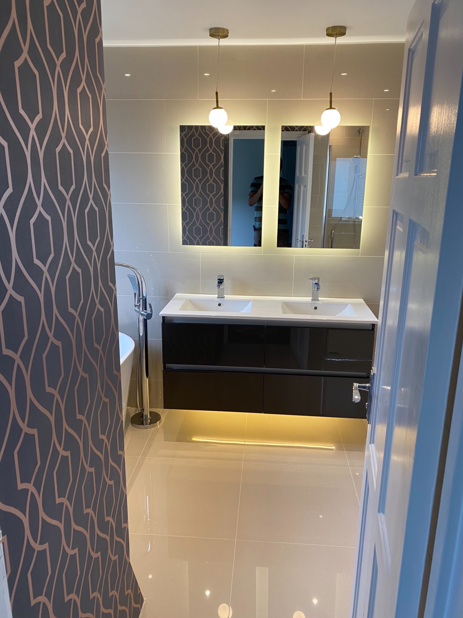 Nowoczesna łazienka z podwójną umywalką na czarnej szafce, podświetlanymi lustrami i złotymi lampami kulami, widok zza ściany z wzorzystą tapetą.
