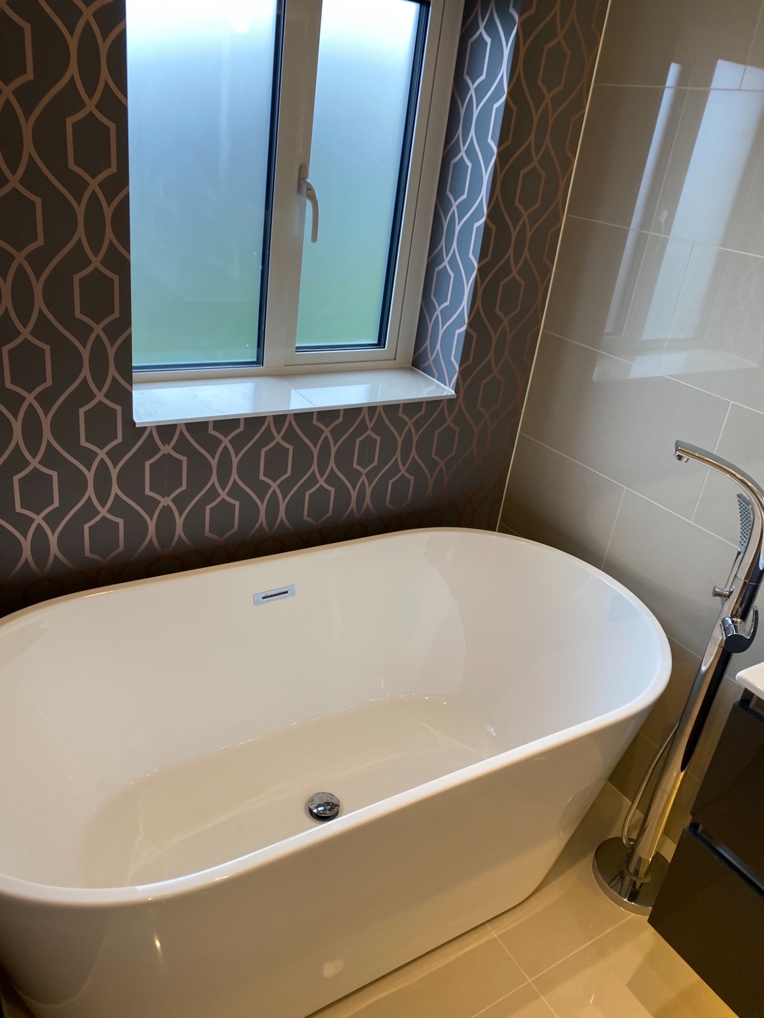 Wolnostojąca biała wanna z chromowaną baterią podłogową, na ścianie tapeta w geometryczny wzór i płytki w odcieniach beżu, okno z mleczną szybą.