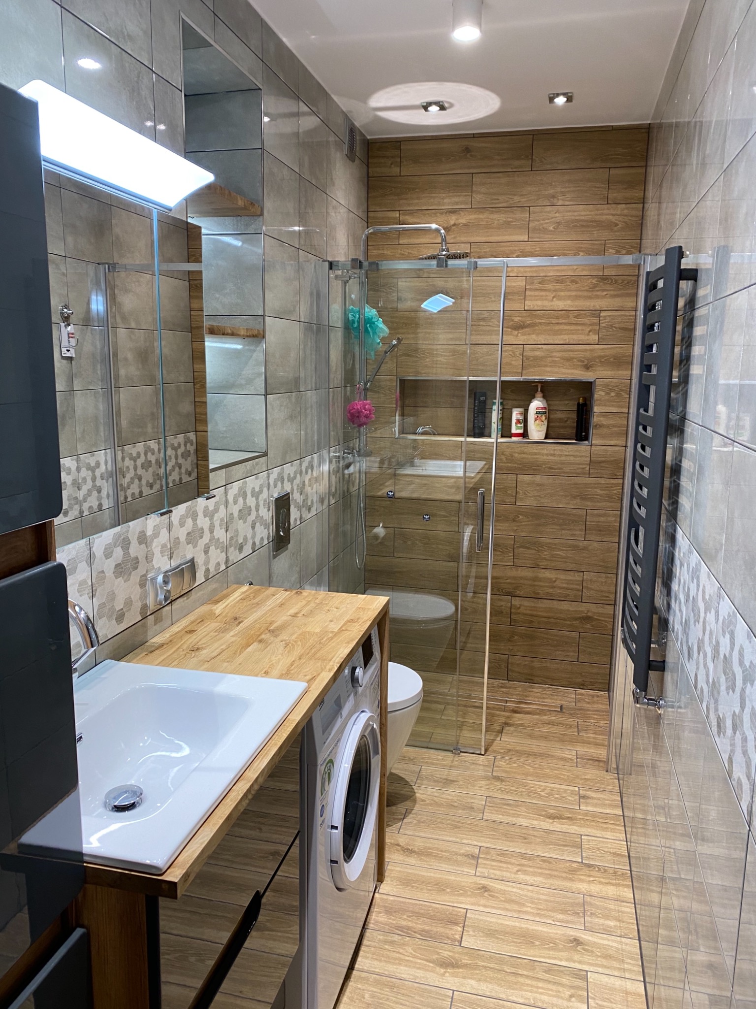 Nowoczesna łazienka z drewnopodobnymi płytkami, przeszkloną kabiną prysznicową, umywalką nablatową na drewnianym blacie i pralką w zabudowie.