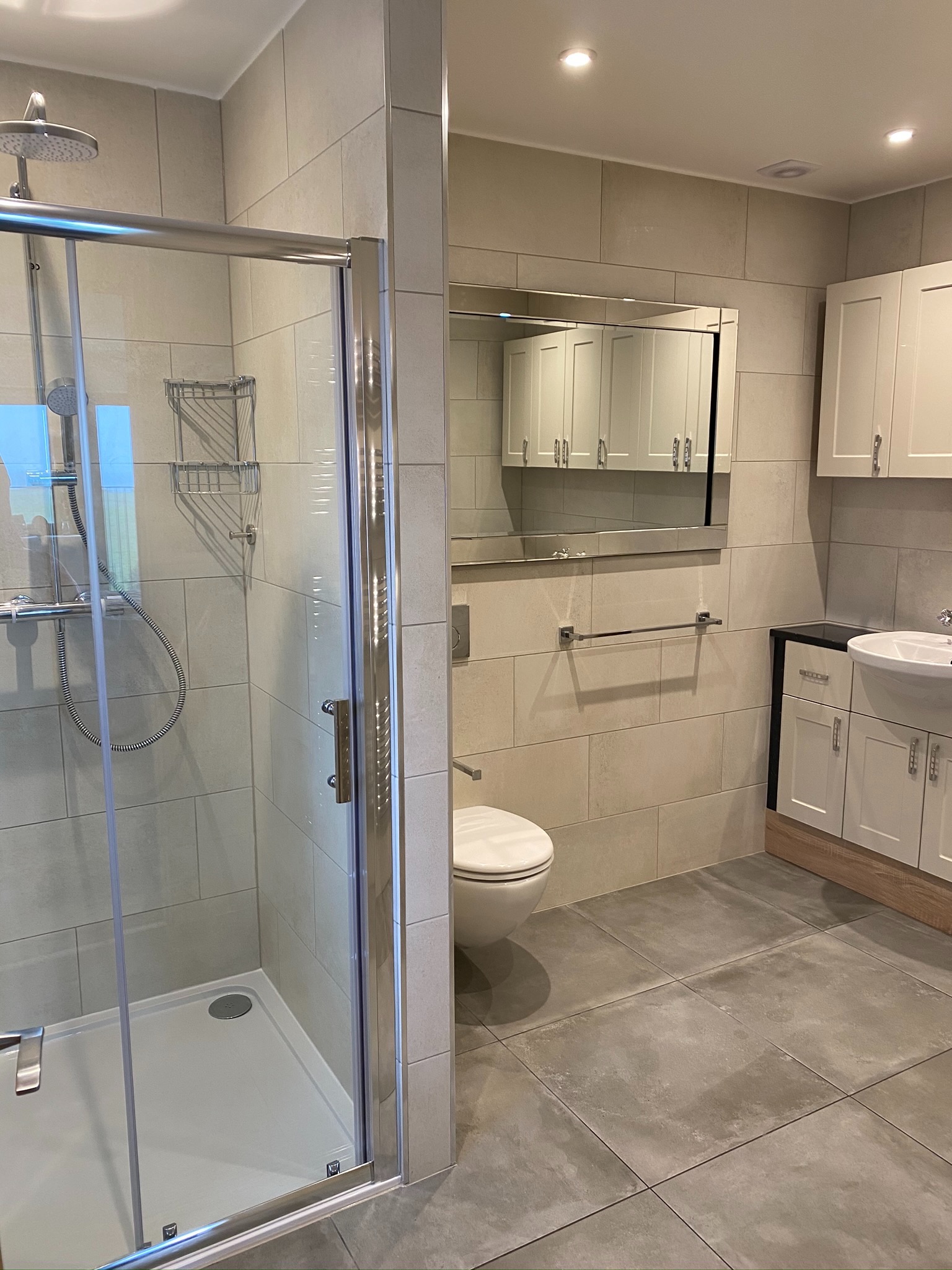 Nowoczesna łazienka z szarą płytką, kabiną prysznicową z chromowanymi detalami, wiszącą toaletą i szafkami w kolorze złamanej bieli z lustrem.