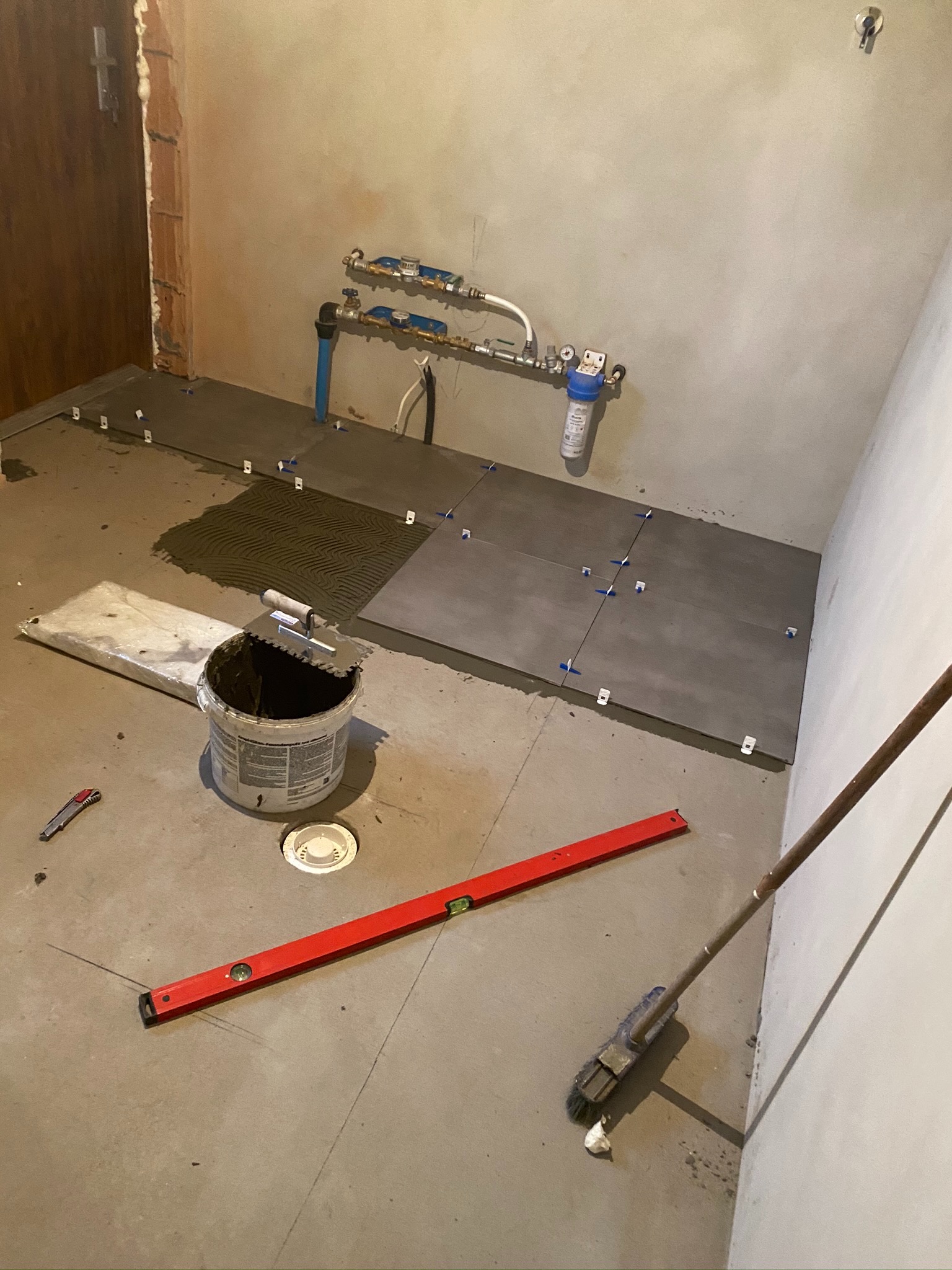 Układanie szarych płytek na podłodze w pomieszczeniu z widocznymi instalacjami wodnymi, poziomica, wiadro z zaprawą i szpachelka zębatą.