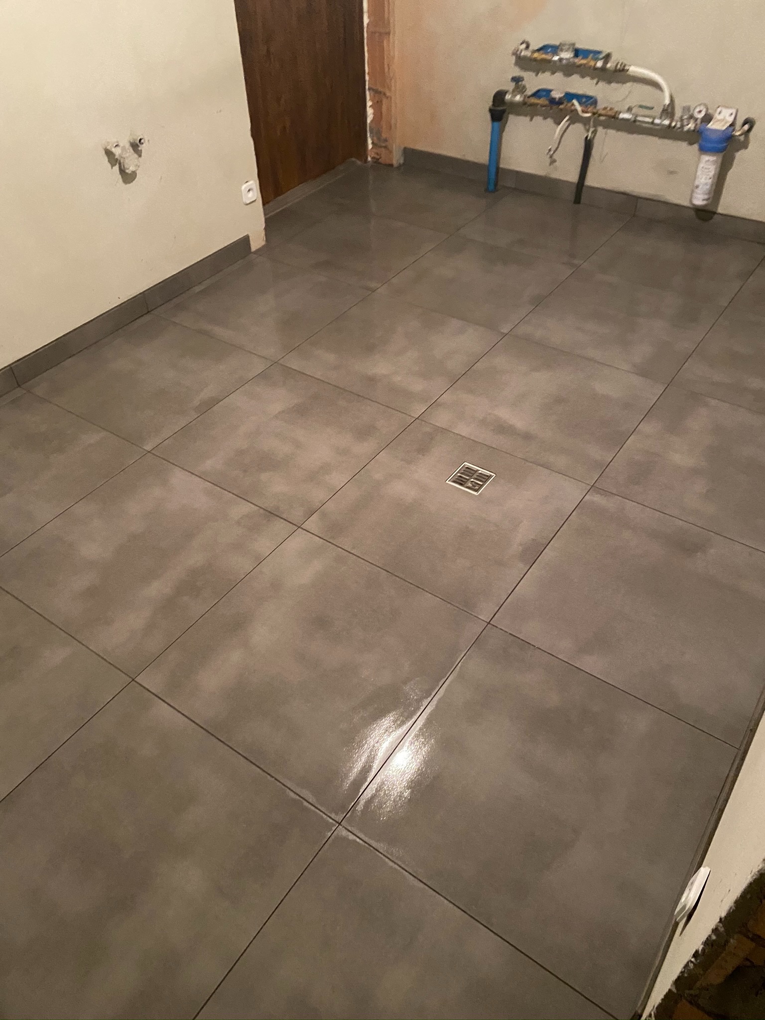 Wykończona podłoga z szarych płytek w łazience z odpływem liniowym, widoczne przyłącza wodne na ścianie i surowy fragment ściany z cegły.