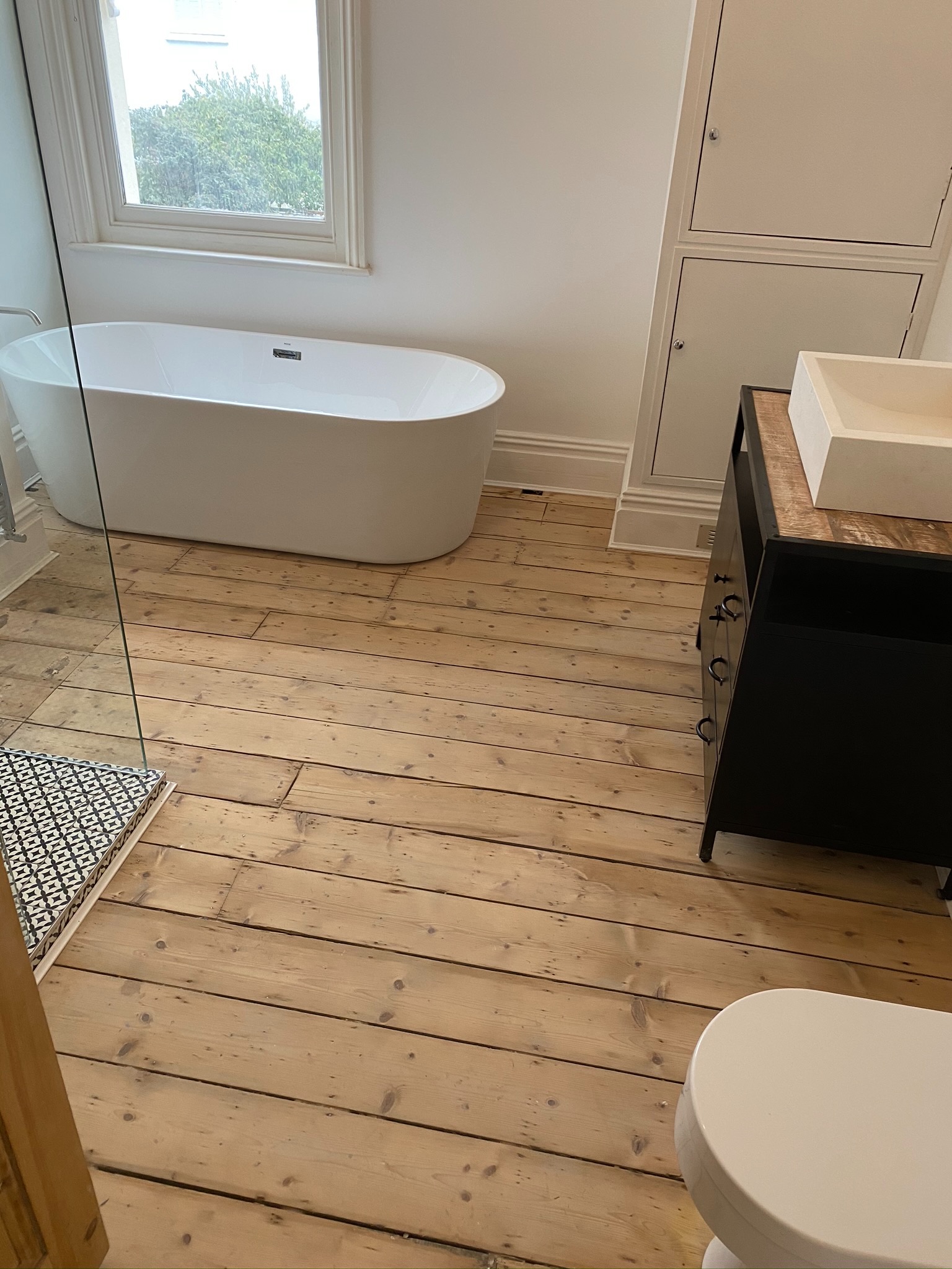 Nowoczesna łazienka z wolnostojącą wanną, drewnianą podłogą i czarną szafką z umywalką na blacie z jasnego drewna.