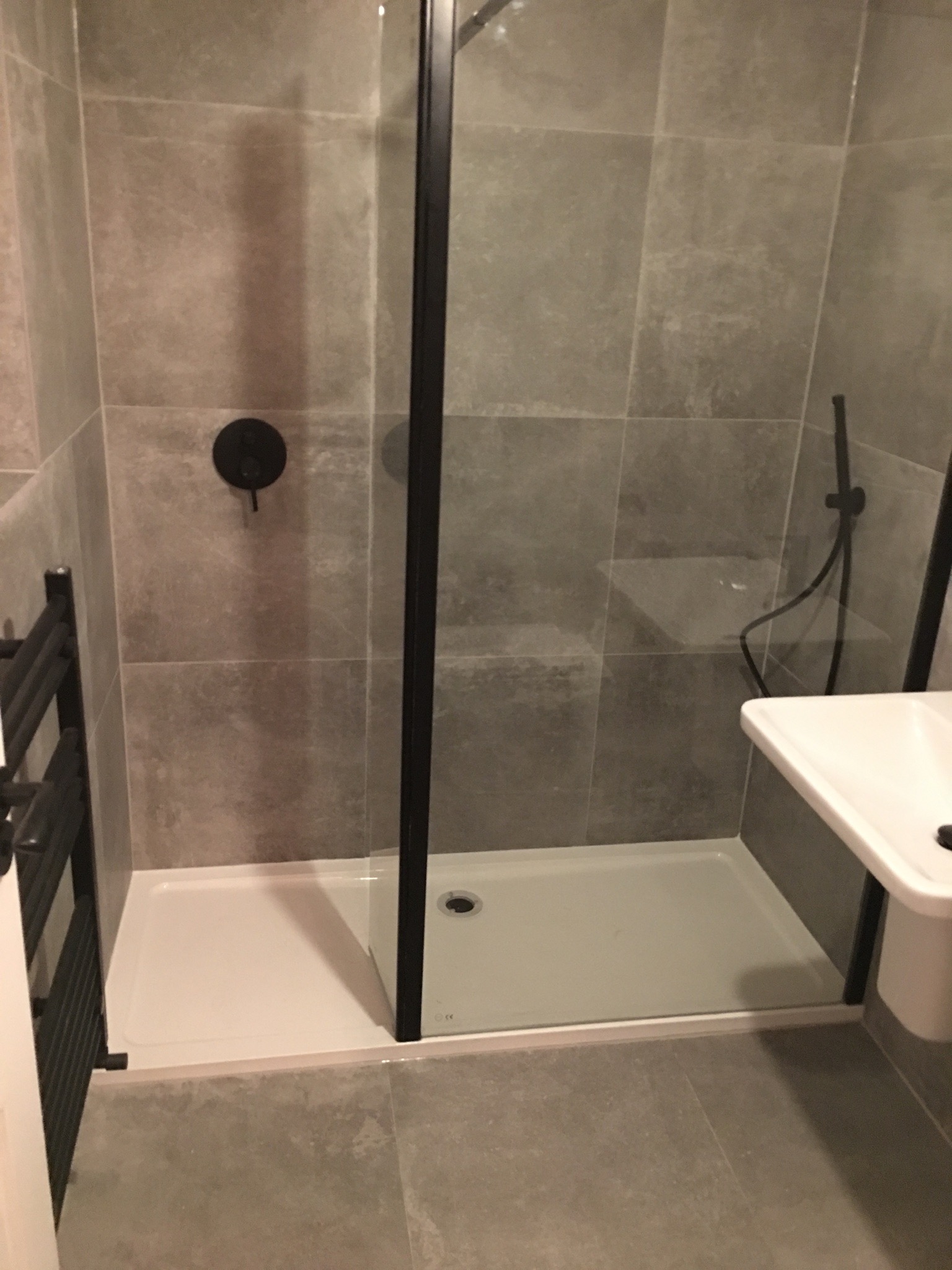 Nowoczesna kabina prysznicowa z czarnymi profilami i szarymi płytkami, widoczna umywalka i grzejnik drabinkowy w łazience.