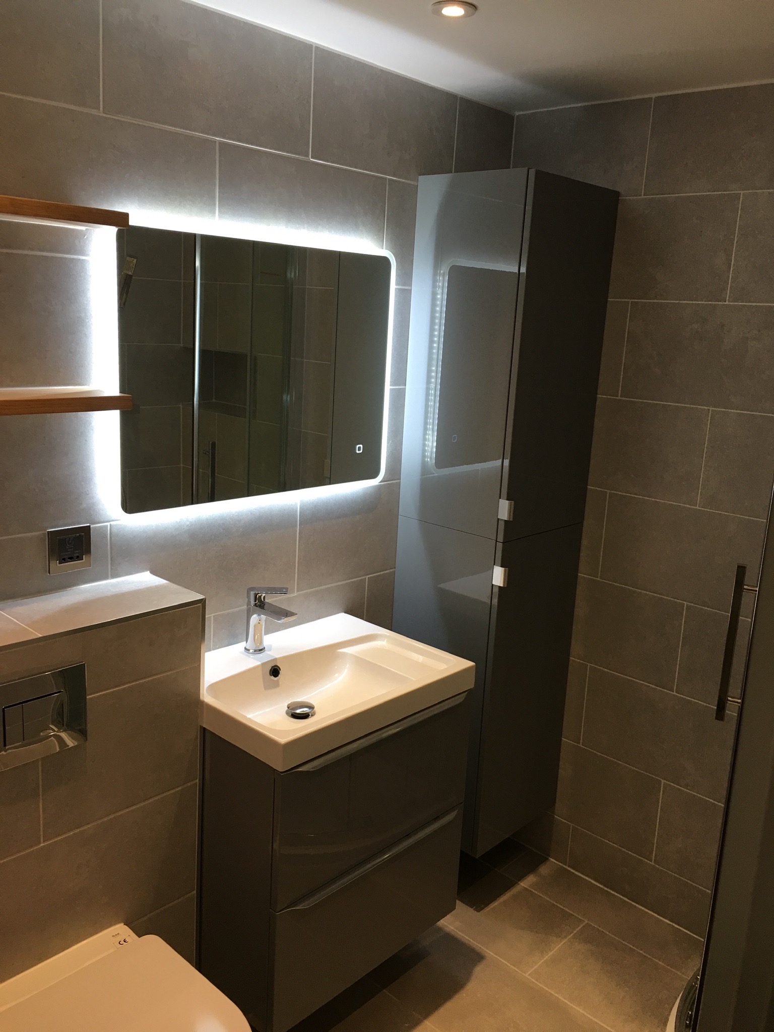 Nowoczesna łazienka w szarościach z podświetlanym lustrem, wiszącą szafką i umywalką nablatową, wykończona płytkami w formacie cegiełki.