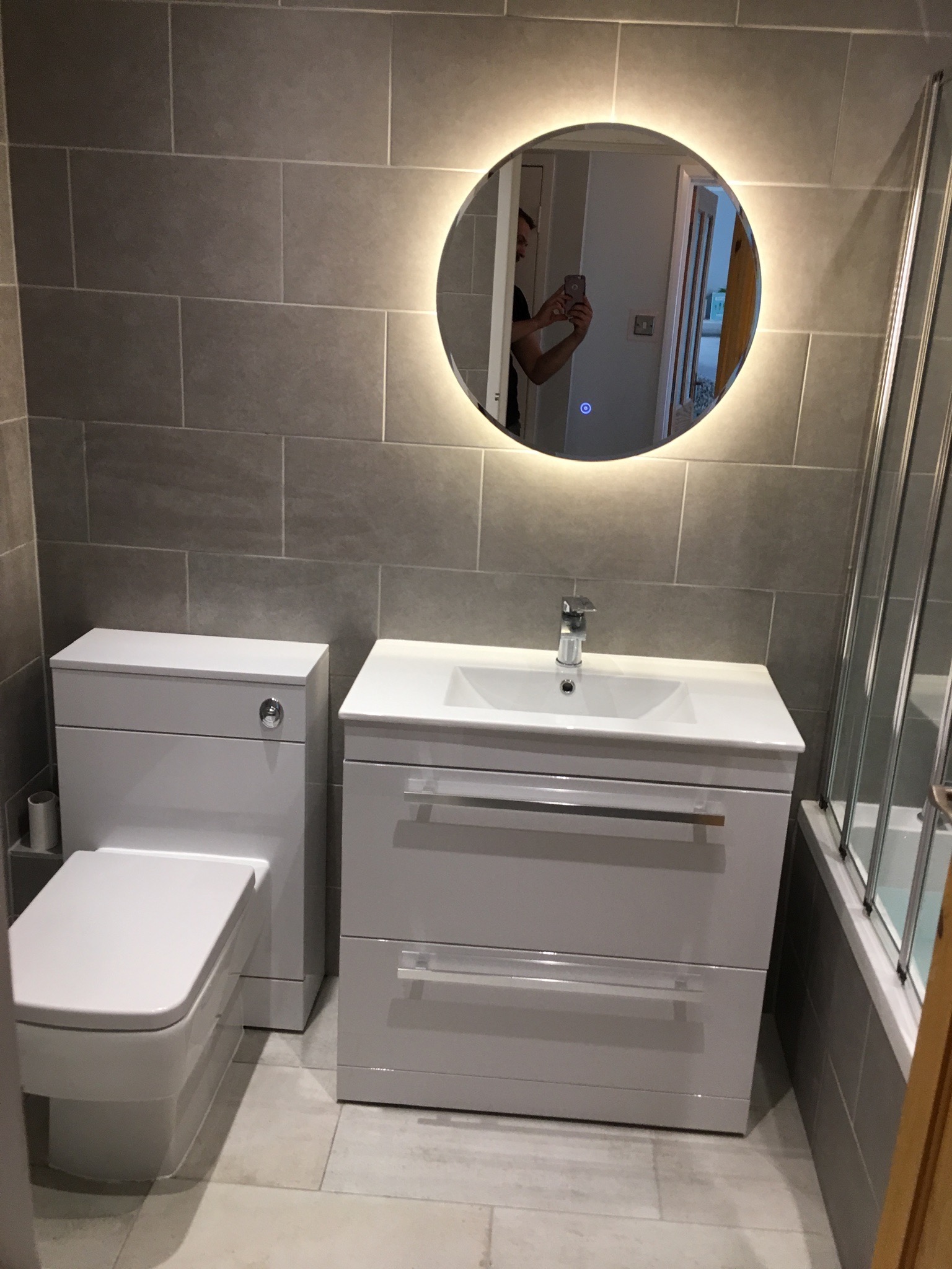 Nowoczesna łazienka z szarą płytką, podświetlanym okrągłym lustrem i białymi meblami łazienkowymi, widoczne odbicie osoby robiącej zdjęcie.