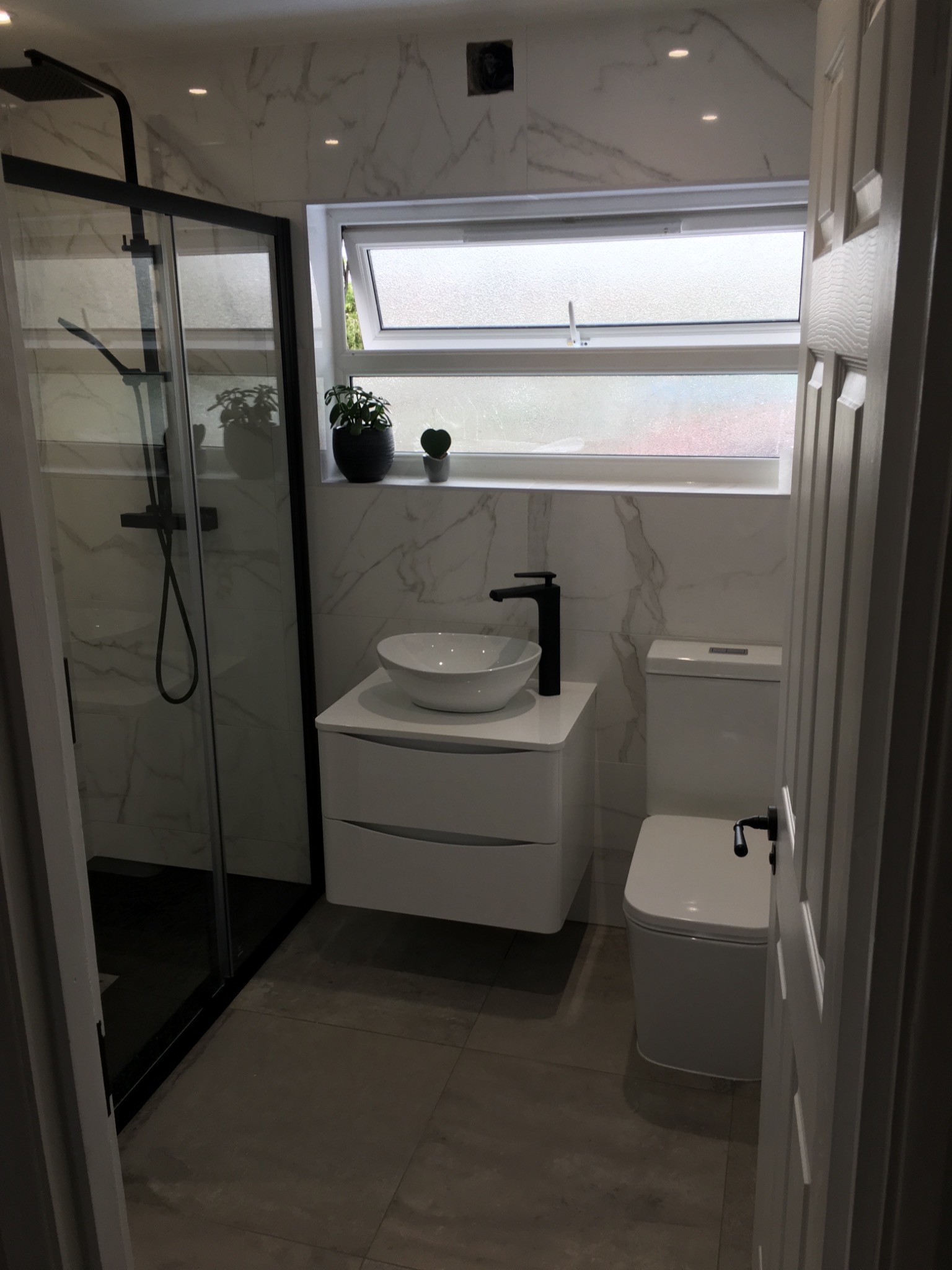 Nowoczesna łazienka po remoncie: kabina prysznicowa z czarną ramą, umywalka nablatowa na białej szafce z szufladami, toaleta i okno z roślinami, wykończenie marmurem.