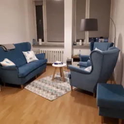 Ustawienie pokoju z niebieską sofą, dwoma fotelami typu uszak i podnóżkiem na wzorzystym dywanie, oświetlone lampą łukową. Za meblami widoczne okno z zasłoniętą roletą.