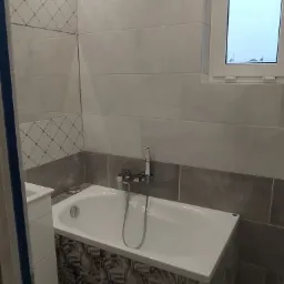 Wykończona łazienka z białą wanną obudowaną płytkami w motywy roślinne, białe płytki ścienne z szarym pasem i dekoracyjną górną częścią, okno w tle.