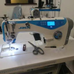 Profesjonalna maszyna do szycia marki Jack z niebieskimi akcentami, nożyczkami, obcinarkami do nitek i kawałkami materiału na blacie roboczym.