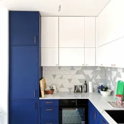 Nowoczesna kuchnia z niebieskimi szafkami dolnymi i białymi górnymi, marmurowy blat, trójkątne płytki na ścianie, chevron na podłodze.