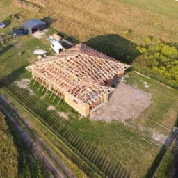 Widok z lotu ptaka na budowę domu jednorodzinnego z widoczną konstrukcją drewnianą dachu, ceglanymi ścianami i otaczającym terenem z elementami rekreacyjnymi.