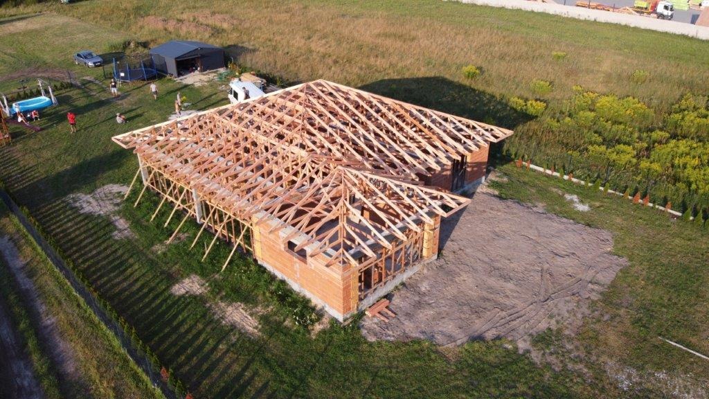 Wznoszenie konstrukcji dachu z drewna na murowanym budynku jednorodzinnym w słoneczny dzień, widok z góry na plac budowy z elementami rekreacyjnymi w tle.